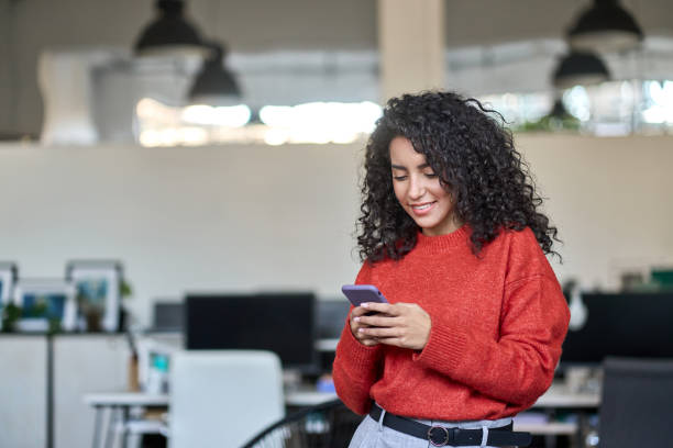 istock Smiling young latin business woman using phone standing in office. 1464209121