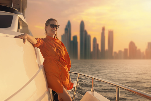 woman in orange dress on luxury yacht deck with Dubai skyline in background at sunset. copy space