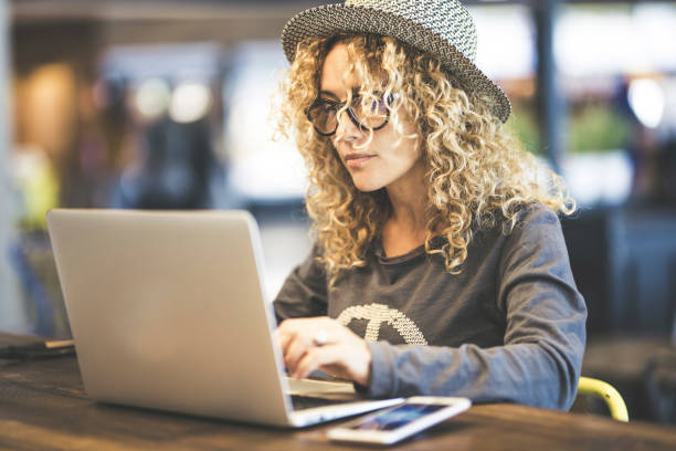 Young female traveler in glasses using laptop communicates on internet with customer in coworking cafe free wireless internet connection place, mobile phone on table. Cozy office workplace remote work stock photo
