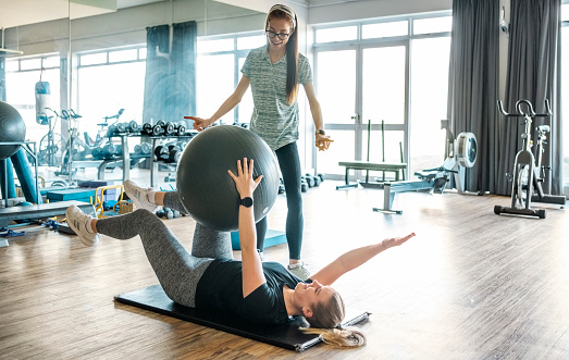 Woman lying on workout mat doing exercises using fitness ball with a biokineticist instructing her at the gym