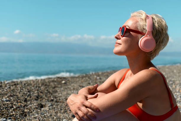 donna sexy bionda in bikini rosso che ascolta musica o podcast in cuffia sulla spiaggia durante le vacanze estive bella giornata calda. - sensuality people mobile phone 30s foto e immagini stock