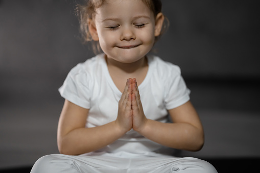 Three years old little girl meditating in a lotus pose on a gray background in dark room. High quality photo