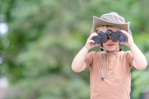 A sweet little boy in a t-shirt and sun hat stands outside with binoculars in his hands as he looks at the world around him.  He has a look of wonder on his face as he looks around his backyard through the tiny lenses.