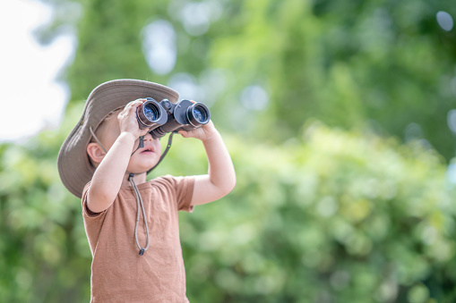 A sweet little boy in a t-shirt and sun hat stands outside with binoculars in his hands as he looks at the world around him.  He has a look of wonder on his face as he looks around his backyard through the tiny lenses.