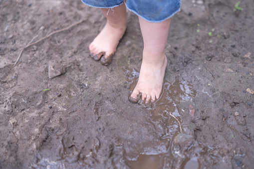 A sweet little boy in shorts and a t-shirt stands in a mud puddle as he jumps and plays in the dirt.  He is barefoot and has a sunhat on as he joyfully splashes about.