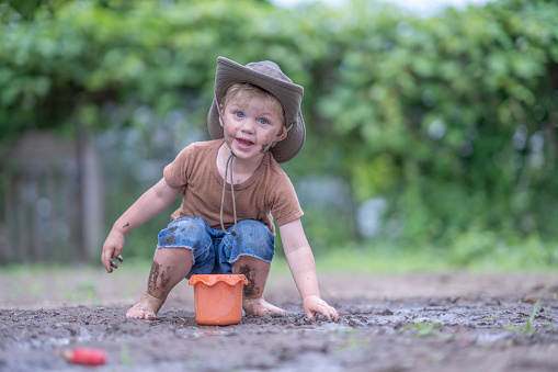 A sweet little boy in shorts and a t-shirt bends down to play in the mud.  He is barefoot and has a sunhat on as he fills his tiny bucket with dirt.