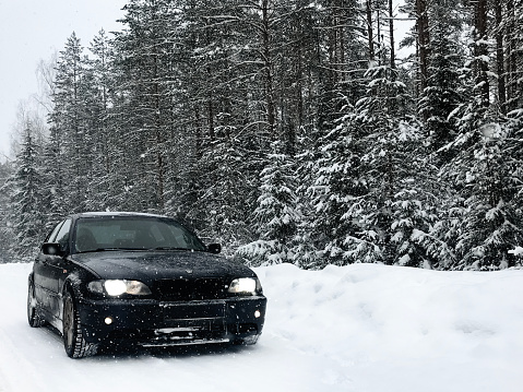 VITEBSK, BELARUS - FEBRUARY 04, 2023: black BMW 3-series E46 with headlights on on a snowy road in a winter forest during a snowfall