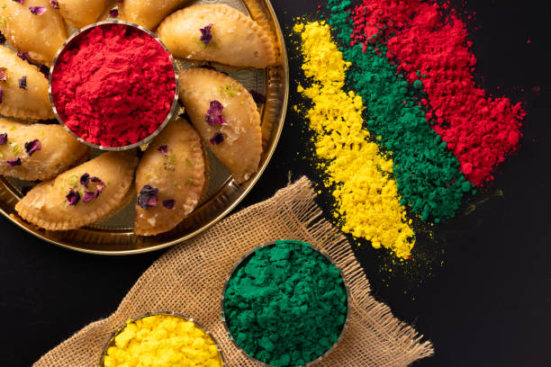 Holi Indian festival of colors celebrating with colorful Gulal Abeer mawa gujia mithai pirukiya sweets presented on a brass plate and scattered isolated on black background stock photo