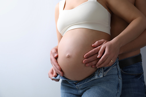 Man touching his pregnant wife's belly on light background, closeup. Space for text