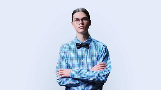 Portrait of confident young male student on white studio background. Serious looking at camera guy teenager in glasses shirt with bow tie, with crossed arms. Youth college university education concept