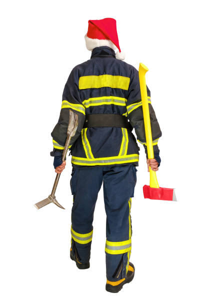 Full body African American fireman in fireproof uniform and red santa hat, holds axe and crowbar in hands over white background isolated. View from the back