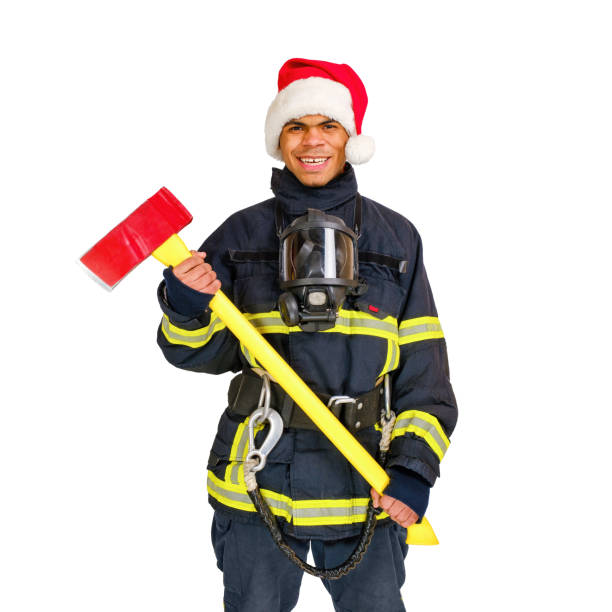 Young African-American fireman in fire-proof uniform and red santa hat, holds axe in hands over white background isolated.