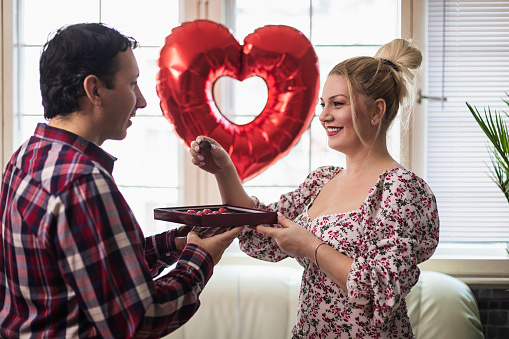 Couple enjoying a romantic moment at home while celebrating Valentine's Day with chocolate candies in hand and air balloons in the shape of a heart in the background