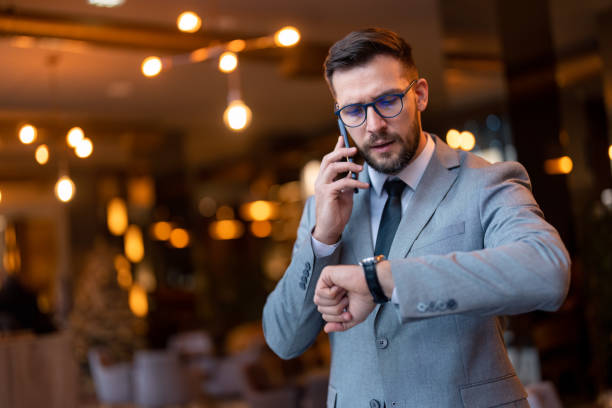 красивый обеспокоенный бизнесмен разговаривает на смартфоне, глядя на ручные часы - checking the time urgency watch business стоковые фото и изображения