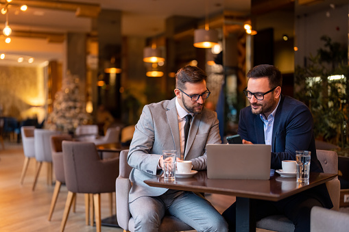 Two businessmen, professional managers, using smartphone and laptop computer in co-working, checking corporate apps while sitting in city coffee shop or restaurant. Stylish men on meeting in coffee bar.