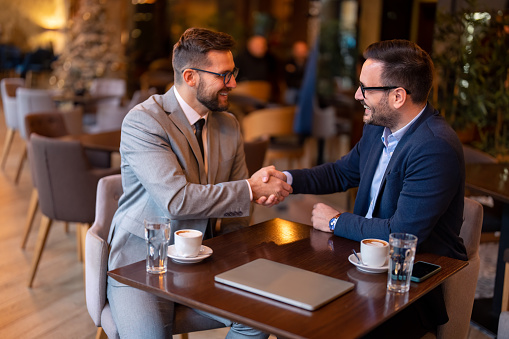 Two smiling confident male business partners at business meeting in restaurant shaking hands as sign that they have reached an agreement, finishing up a meeting or setting goals, sharing productive ideas.