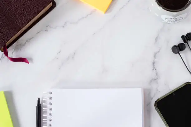 Blank notebook, holy bible book, earphones, smartphone, paper notes, and cup of coffee on white marble background. Top table view. Copy space for text. Christian study, biblical education concept.