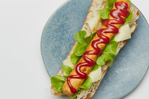 Solo hotdog sandwich on a handmade ceramic plate, made of fresh baguette, lettuce, ham, cheese, Spanish onion, tomato, mayo and ketchup. Isolated on a white background, top view image with a large copy space area
