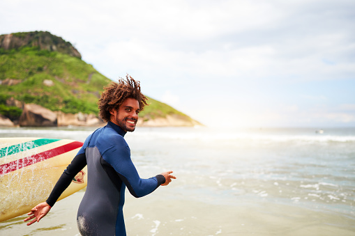 young mixed race brazilian man in surf suit walking along the beach shore towards the sea smiling happy