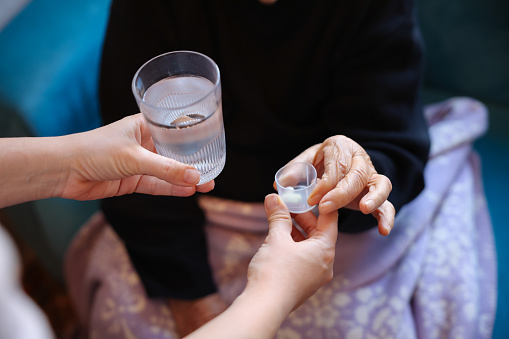 An older man and a young girl at home. The granddaughter standing nearby and giving medicine and a glass of water to the grandfather who is sitting on the sofa