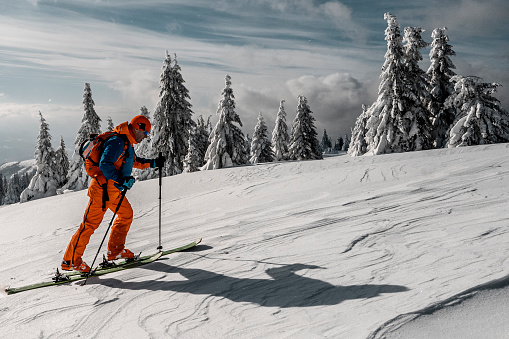 A  male adventurer with proper winter clothes, skis, boots, poles and all other details seen ski touring in the mountains surrounded by mesmerizing nature during a winter vacation.