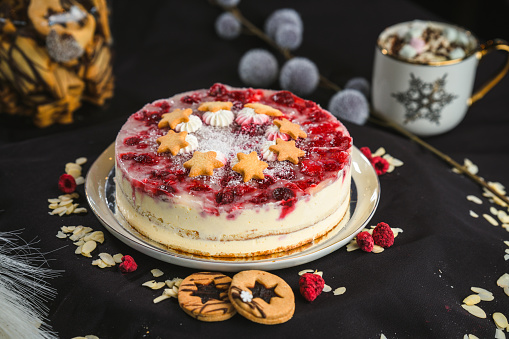 High angle view of a round  raspberry cheese cake presented in front of a black studio background, raspberries and almond flakes scattered in the foreground, cookies next to the plate. A cup of hot chocolate with marshmallows in the background. Studio shot, full length image.