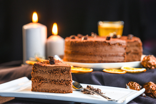 A slice of made to order    chocolate cake on a white plate, spoon and chocolate shavings next to it, a whole round cake in the background, everything festively decorated with Christmas decoration: dried lemon slices, a pine cone and lit candles. Studio shot, full length image. Focus on the slice, side view.