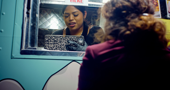 A young Latina woman working in a taco truck on a city street at night takes an order from a customer with her back to the camera.