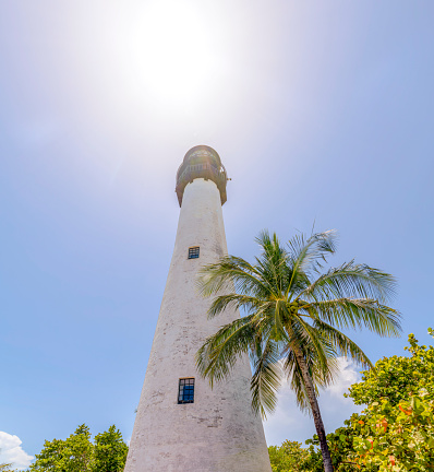 Low angle view of Cape Florida Lighthouse at Bill Baggs Cape Florida State Park, Miami, Florida. Tall lighthouse tower with trees outside below the sun in the clear sky.