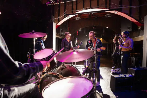 A drummer playing onstage with a rock band during the sound check before a live show at a small venue. A photographer is taking pictures of them.