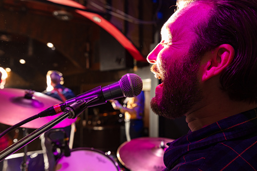 Close up on the face of a man performing on stage with his band at a small venue, playing drums and singing backing vocals.