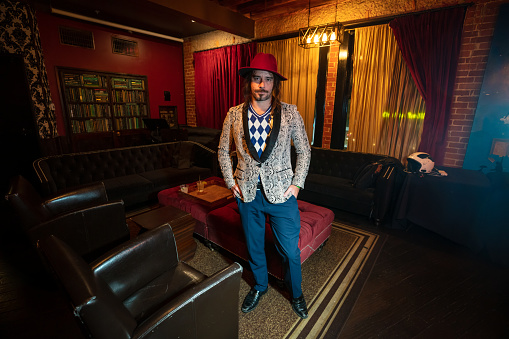 Portrait of a snappily dressed man wearing a red hat standing in the backstage area of a music venue, a large room decorated with ornate wallpaper.