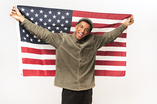 Happy latin man fighter for rights holding wide open United States flag behind back, smiling and cheerful guy protesting against racism, isolated on white wall