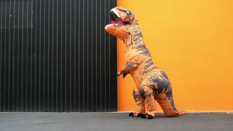 One happy and funny dinosaur costume dancing in the street with a orange colorful background - t-rex having fun - funny man inside of a costume of dino