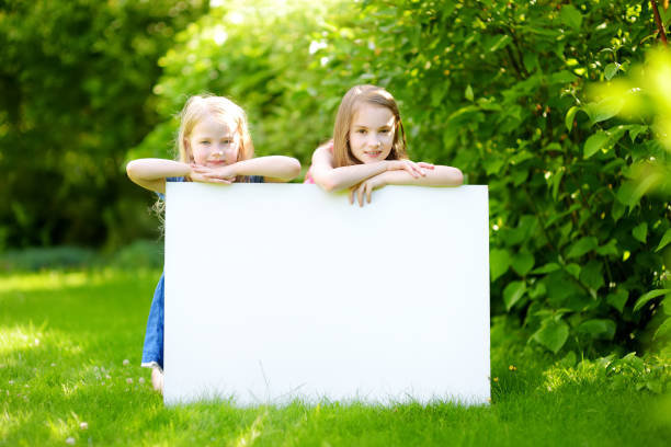 two cute little sisters holding big blank whiteboard on sunny summer day outdoors - preschooler childhood outdoors cheerful imagens e fotografias de stock