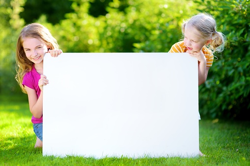 Two cute little sisters holding big blank whiteboard on warm and sunny summer day outdoors