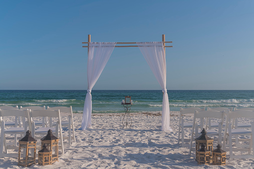 Beach wedding aisle with white curtain arch and lantern decorations near chairs in Destin, Florida. Simple wedding ceremony aisle on a white sand beach with views of the blue ocean waves and sky.