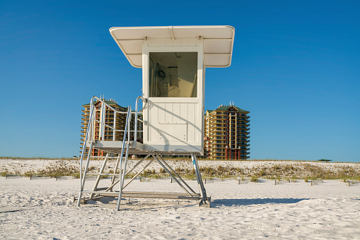 Lifeguard tower with glass panel on a white sand beach against the hotel building at Destinm, FL. There are fence barriers from sand dunes near the hotel at the back of the lifeguard tower.