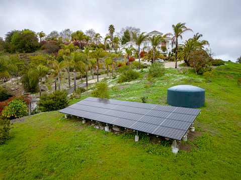 Fallbrook, California- Solar panels and cistern tank near the driveway of a mansion in a farm. There is a driveway heading to a mansion at the back with palm trees outdoors against the cloudy sky.
