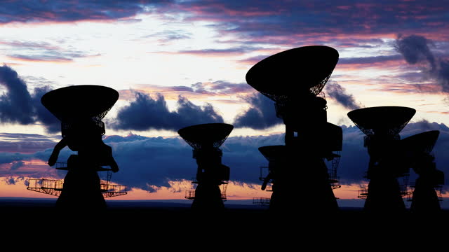 Silhouettes of huge radio telescopes facing up in the sky with the colored sunset sky in the background