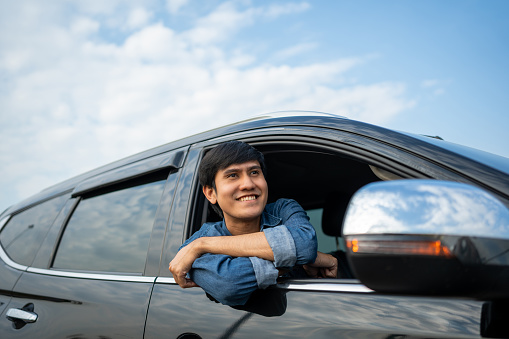 A young Asian man drives a car on a clear day. With beautiful blue sky. He smiling driving to travel by car. Sticking her head outta the windshield