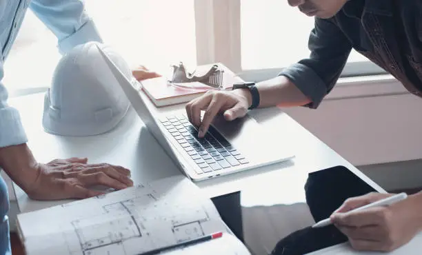 Photo of Two  architect colleagues discussing data working on digital tablet and laptop computer with architectural project at construction site on desk at office