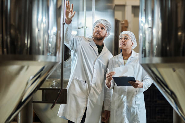 deux travailleurs portant des blouses de laboratoire inspectent l’équipement d’une usine alimentaire - usine agro alimentaire photos et images de collection
