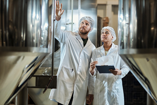 Waist up portrait of two workers wearing lab coats inspecting equipment at food factory