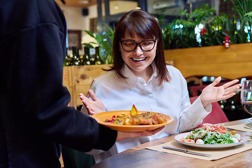 Middle-aged smiling woman sitting at table in restaurant with plate of salad on table, rejoices of cooked fish food in hands of waiter. Delicious food, nutrition, leisure lifestyle, 40s people concept
