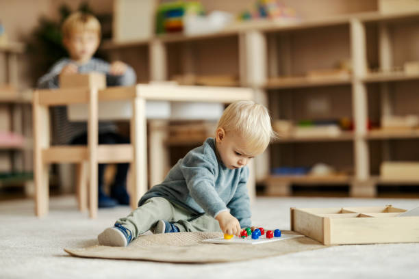 A 1 year old girl crawls on the carpet while smiling. A group of babies sitting next to her look around curiously. They are in a colorful daycare environment.