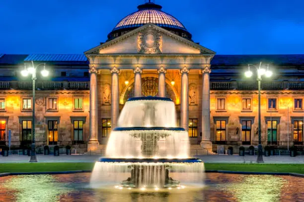 The facade of a neo-Renaissance building in the evening with artificial lighting and the illuminated cascades of a fountain in the foreground in a publicly accessible square
