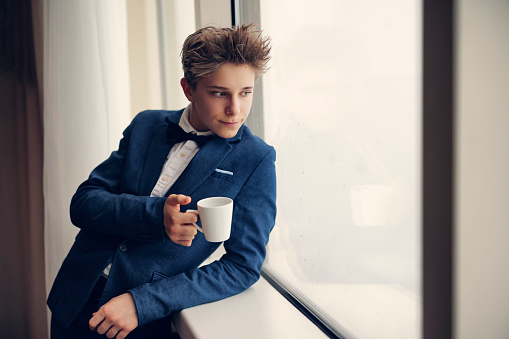 Portrait of a cute teenage boy wearing a suit and a bow tie. The boy is looking at the city and holding a cup of coffee.\nCanon R5