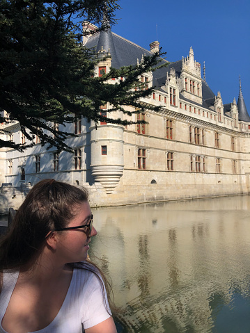 Vertical closeup photo of a young woman, in profile view,  looking over the reflections on theMirror Lake next to the French Renaissance castle at Azay-le-Rideau.\nLoire Valley, Maine et Loire Department, France. 31st March, 2019.