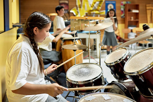 Side view of 11 year old in braids smiling as she plays with background classmates on congas, bongos, and flute.